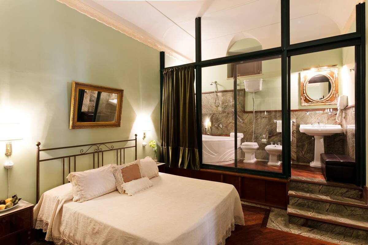 A hotel room with a bed, a bathroom visible through a glass door, and a framed picture on the wall.