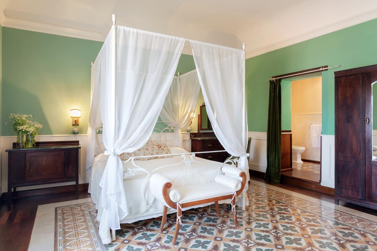 A four-poster canopy bed with a wooden frame and white bedding, a wooden nightstand, a patterned floor, and a green wall.