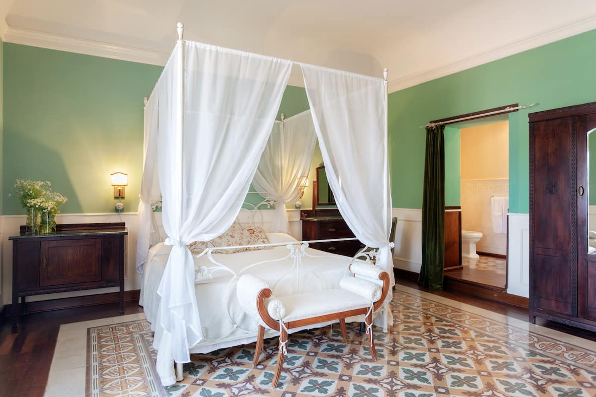 A four-poster canopy bed with white linens, a wooden bench, and a patterned floor.