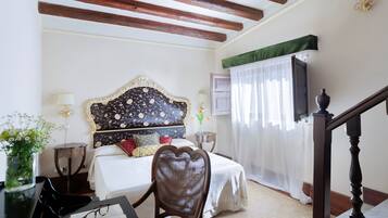 Quarto com teto de madeira, cama com cabeceira florida, escrivaninha com um vaso de flores e uma cadeira.
