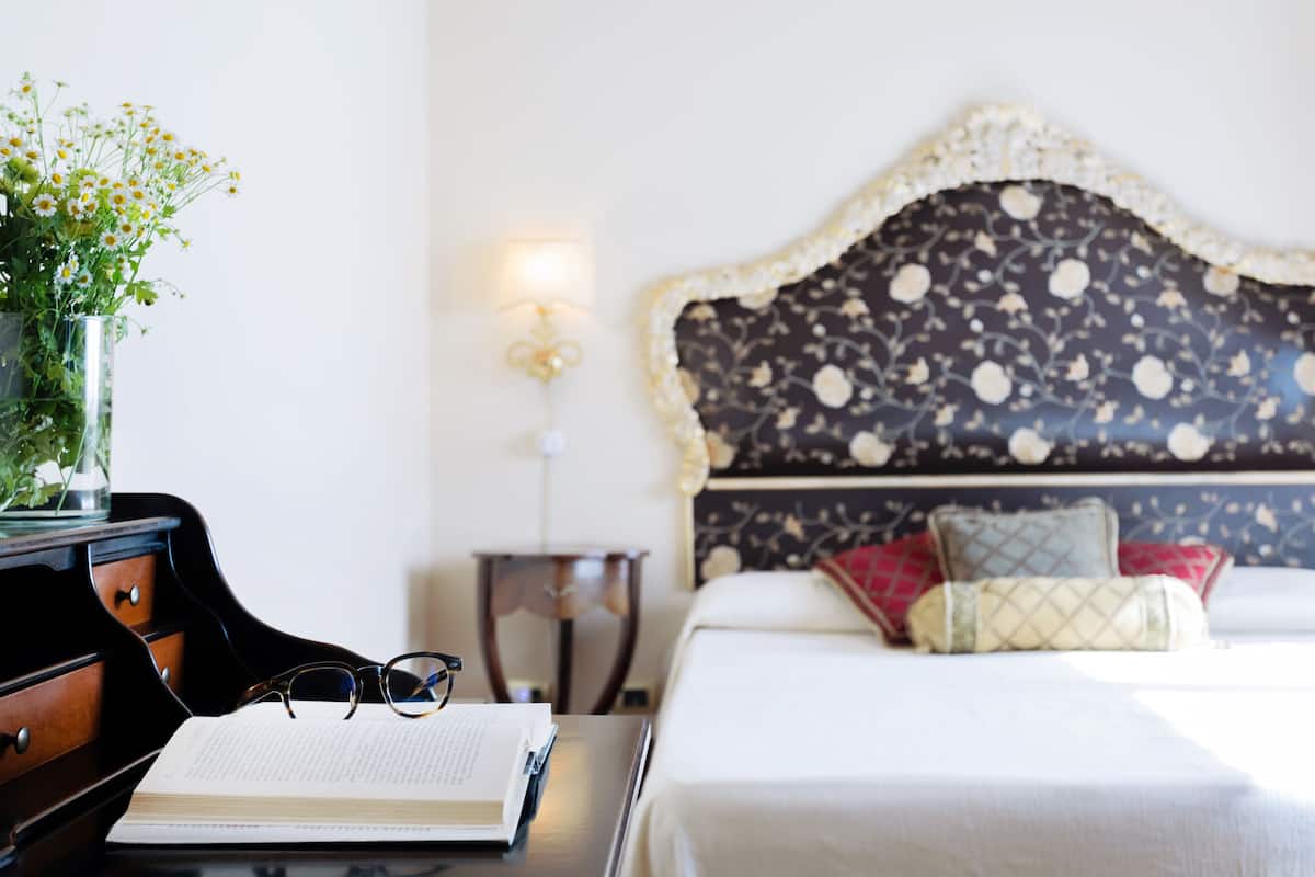A neatly made bed with a floral headboard, a bedside table with a book and glasses, and a vase with flowers.