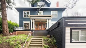 A two-story house with a front porch, a staircase leading to the front door, and a small garden area. - A pied-à-terre near bistros, shops, and sports stadium w/ parking and EV charger (Portland)