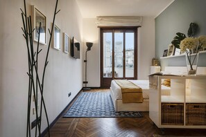A room with a bed, a bedside table, a lamp, a window with a view of buildings, and a wall with framed pictures.