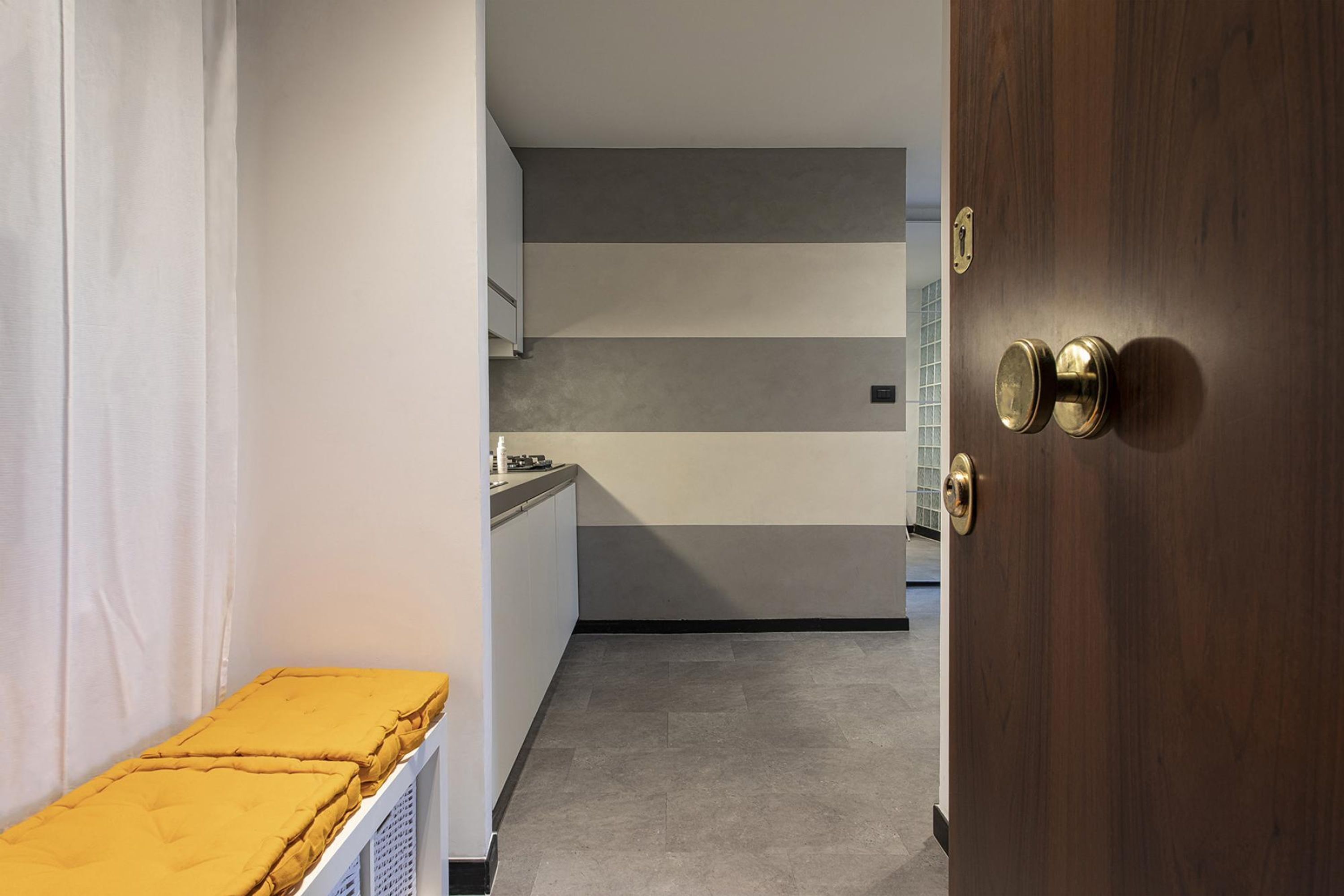 A narrow hallway with a wooden door on the right, a white radiator on the left, and a kitchen area in the background with grey and white walls.