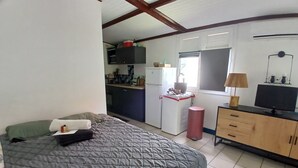 Ein Schlafzimmer mit Bett, Kommode, Fernseher und einer kleinen Küchenzeile im Hintergrund.