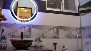 Une salle de bain avec un miroir rond, un lavabo agrémenté d’un récipient décoratif, une plante en pot, un distributeur de savon liquide et un distributeur de serviettes.