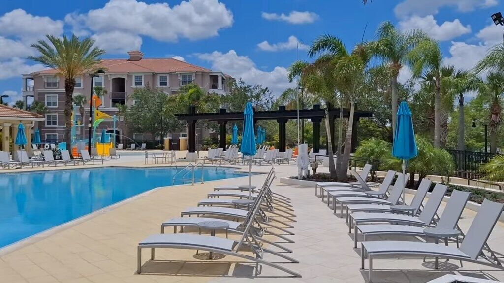 Un espace aménagé au bord de la piscine, avec des chaises longues et des parasols, une piscine à l’eau d’un bleu limpide, et un bâtiment avec un toit rouge en arrière-plan.