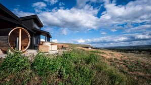 Una casa moderna con un gran ventanal de madera, rodeada de un paisaje cubierto de hierba y un cielo azul despejado.