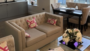 Een moderne woonkamer met een bank, een glazen salontafel en een eethoek met een tafel en stoelen. Achtergrond is een keuken met kastjes en een spoelbak.