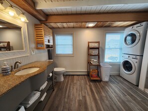 A bathroom with a wooden floor, two sinks, a toilet, a washing machine, and a shelf with towels. - Hampton Beach ♡ Ocean Retreat - Steps From The Sand and Strip! (Hampton)