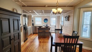 A kitchen with wooden floors, a dining table with chairs, a chandelier, and a view of the outside through a window. - Hampton Beach ♡ Ocean Retreat - Steps From The Sand and Strip! (Hampton)