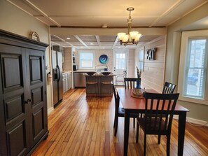 A kitchen with wooden floors, a dining table with chairs, a chandelier, and a view of the outside through a window. - Hampton Beach ♡ Ocean Retreat - Steps From The Sand and Strip! (Hampton)
