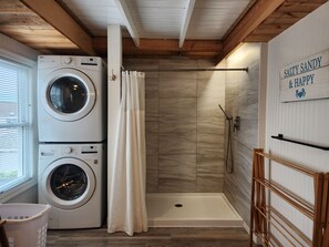 A laundry area with two stacked washers and dryers, a shower with a wooden door, and a sign on the wall. - Hampton Beach ♡ Ocean Retreat - Steps From The Sand and Strip! (Hampton)
