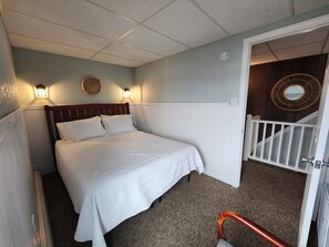 A hotel room with a bed, two wall-mounted lights, a round mirror, a white door, and a white railing. - Hampton Beach ♡ Ocean Retreat - Steps From The Sand and Strip! (Hampton)