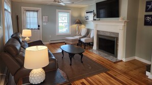 A living room with a sofa, chairs, a fireplace, a television, and a ceiling fan. - Hampton Beach ♡ Ocean Retreat - Steps From The Sand and Strip! (Hampton)
