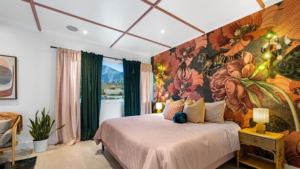 A bedroom with a large bed, a nightstand with a lamp, a potted plant, and a view of mountains through the window.