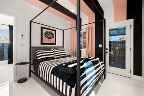 A modern bedroom with a four-poster bed, a framed artwork above the bed, and a view of the outdoors through a glass door.