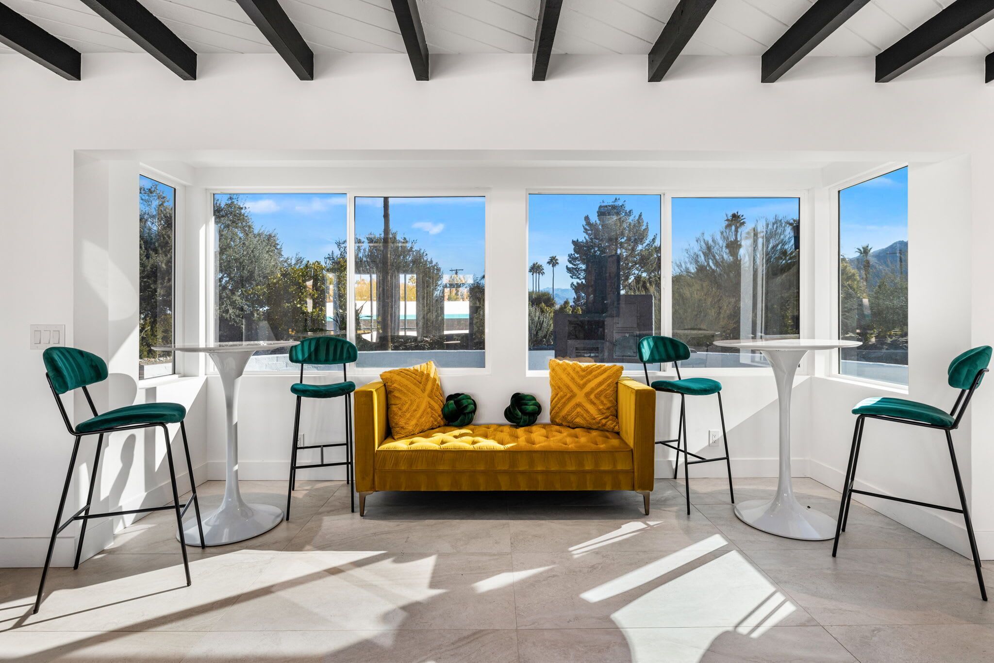 Uma sala de estar moderna com um sofá amarelo mostarda, cadeiras verdes e grandes janelas com vista para árvores e montanhas.