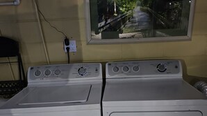 A laundry room with two white front-loading washers and dryers, a window with a view of a bridge, and a wall-mounted electrical outlet.