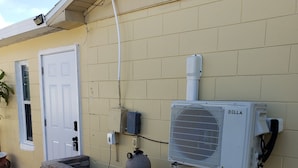 An outdoor air conditioning unit mounted on a yellow wall, a brick planter with a potted plant, and a white door.