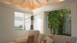 A cozy room with a beige sofa, striped cushions, and a decorative pillow. There are two windows with blinds, one of which has a view of greenery outside. - Villa Douceur Caraïbes, 500m from the beach, pool under the palm trees (Saint-Francois)