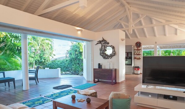 A spacious living area with a large TV, wooden furniture, and a view of lush greenery outside.