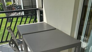 Un balcon avec une table et des chaises grises, une porte vitrée et une vue sur un jardin.