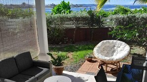 En täckt uteplats med en soffa i rotting, ett runt vitt bord och en stol. Utsikten sträcker sig över havet och i bakgrunden syns palmer.