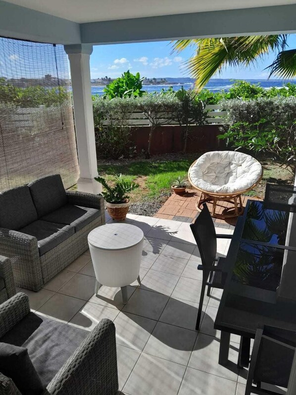 A covered patio with a wicker sofa, a round white table, and a chair. There is a view of the ocean and palm trees in the background. - Apartment Seaside sea view and direct beach access (Le Moule)