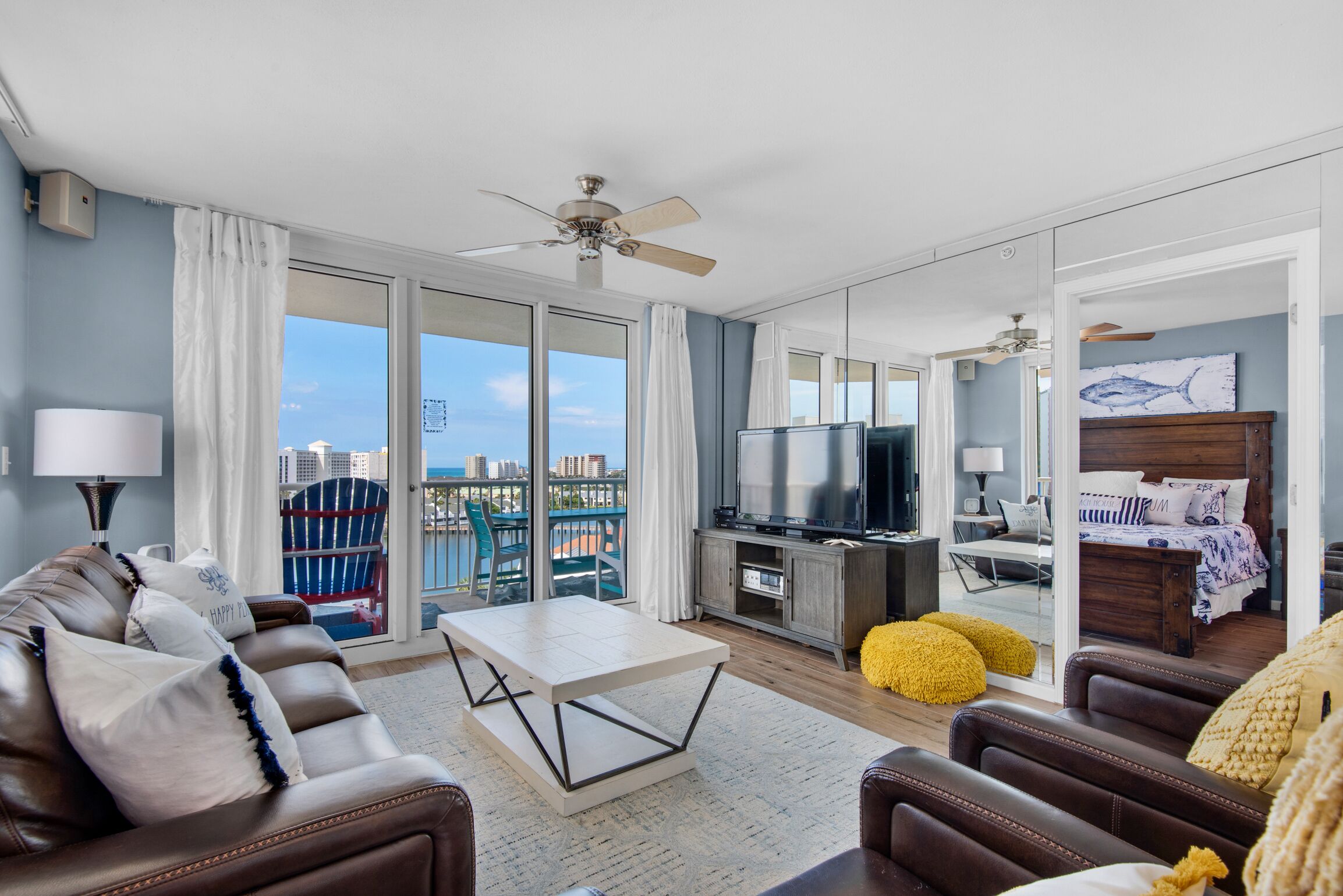 A modern living room with a sofa, coffee table, and TV, a balcony with a view, and a bedroom with a bed and artwork.