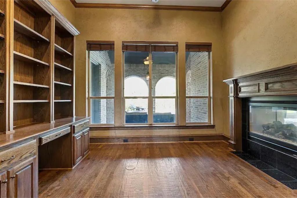 A room with hardwood flooring, built-in wooden shelving, a fireplace, and large windows with a view of brick buildings.