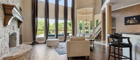 A spacious living room with a stone fireplace, large windows, a staircase, and a bar area.