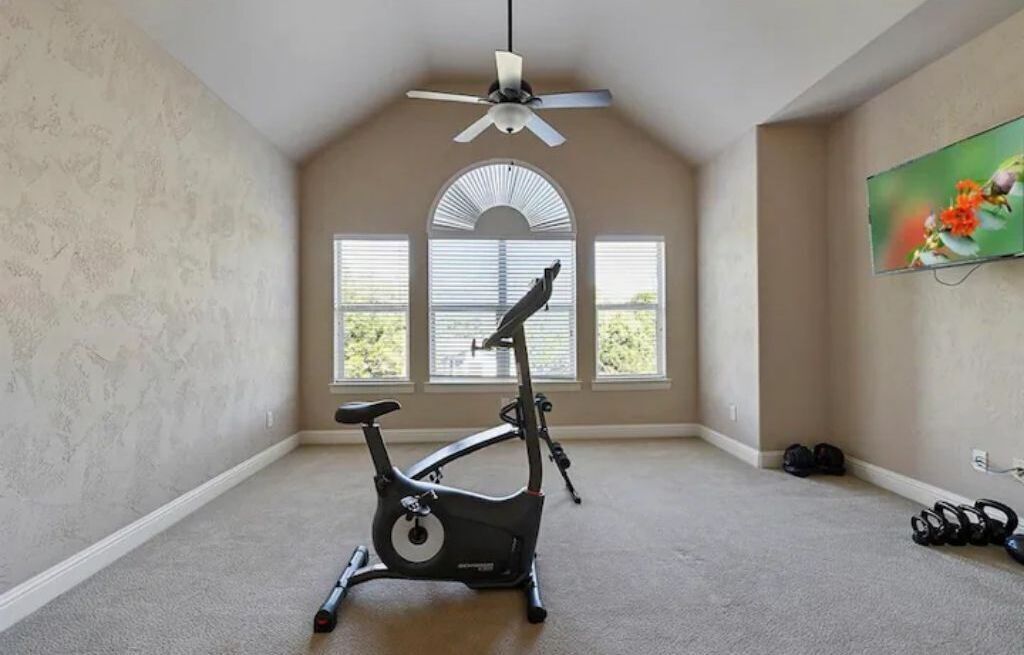 Un'ampia stanza con ventilatore a soffitto, ampie finestre con tende e una cyclette, con un televisore montato a parete.