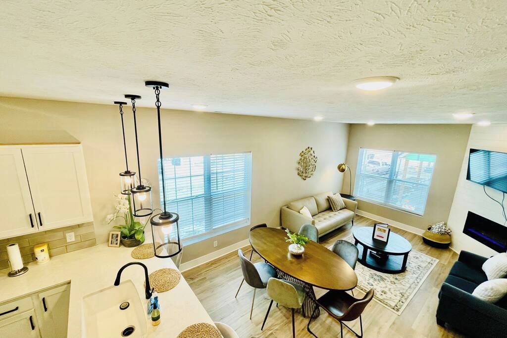 A modern kitchen with white cabinets, a dining area with a wooden table and chairs, a sofa, and a TV.