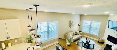 A modern kitchen with white cabinets, a dining area with a wooden table and chairs, a sofa, and a TV.