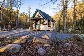 Une cabane au toit de chaume, entourée d’une allée de gravier et d’une zone boisée.