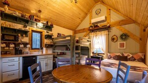 A cozy cabin interior with a dining area, bunk beds, wooden beams, and a kitchen with shelves. - Storybook Cottage @ Four Fillies Lodge-Glamping (Peterstown)