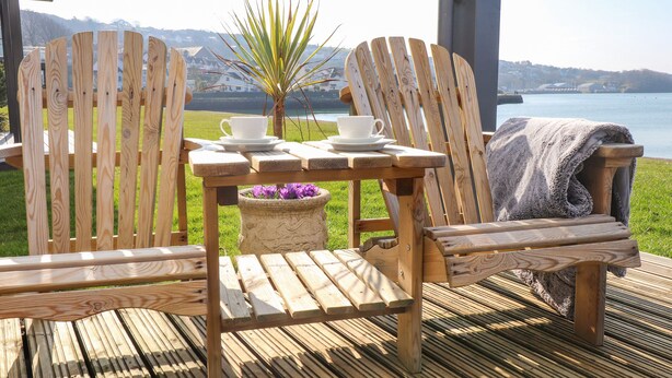 Une terrasse en bois avec deux fauteuils Adirondack, une table avec une tasse et une soucoupe, et une plante en pot.