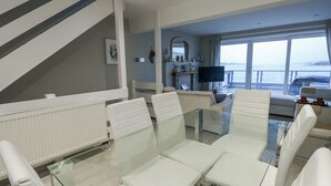 Un salon moderne avec une table à manger en verre, des fauteuils en cuir blanc, une grande fenêtre donnant sur la mer et un téléviseur à écran plat.