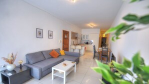 A modern living room with a grey sofa, a white coffee table, and a small side table with a vase. There is a kitchen in the background with white cabinets and a dining area. - Large and modern 2bedroom with balcony GLUB1-1 (Gzira)