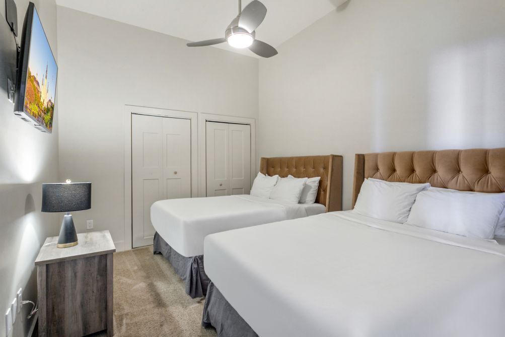 Habitación de hotel con dos camas, ventilador de techo, televisión montada en la pared y mesita de noche con lámpara.