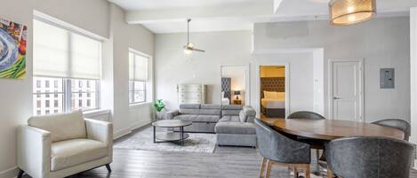 A spacious living room with a gray sectional sofa, a round coffee table, and a dining table with chairs. There are large windows providing natural light, and a ceiling fan is visible.