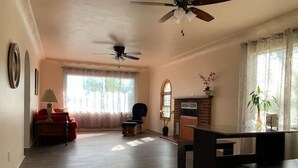 Ein geräumiges Wohnzimmer mit Kamin, Deckenventilator und einem großen Fenster mit durchsichtigen Vorhängen.