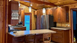 Interieur van een blokhut met een keuken voorzien van houten kastjes, een roestvrijstalen koelkast en een eettafel met stoelen. Er is een bord met huisregels en een uitzicht op de omgeving.