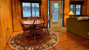 Een gezellig interieur van een blokhut met een eethoek, een bank en uitzicht op een besneeuwd landschap door de deur.