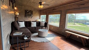 Un salon chaleureux avec un canapé d’angle, un pouf et une petite table basse. On y trouve un ventilateur de plafond, une lampe et une vue sur l’extérieur grâce à de grandes fenêtres.