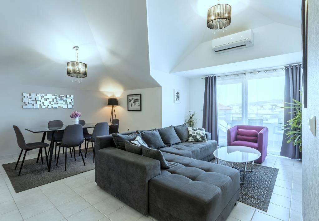 A modern living room with a grey sectional sofa, a round coffee table, and a purple armchair. There is a dining area with a table and chairs, and a large window offering a view of the city.