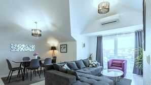 A modern living room with a grey sectional sofa, a round coffee table, and a purple armchair. There is a dining area with a table and chairs, and a large window offering a view of the city. - Apartment in Kaštel Novi with Balcony, Air condition, WIFI, Washing machine (5105-1) (Kaštel Novi)