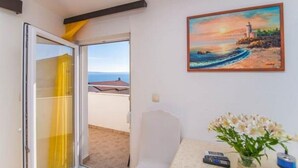 A room with a painting of a lighthouse, a small table with a vase of flowers, and a view of the sea through a glass door.