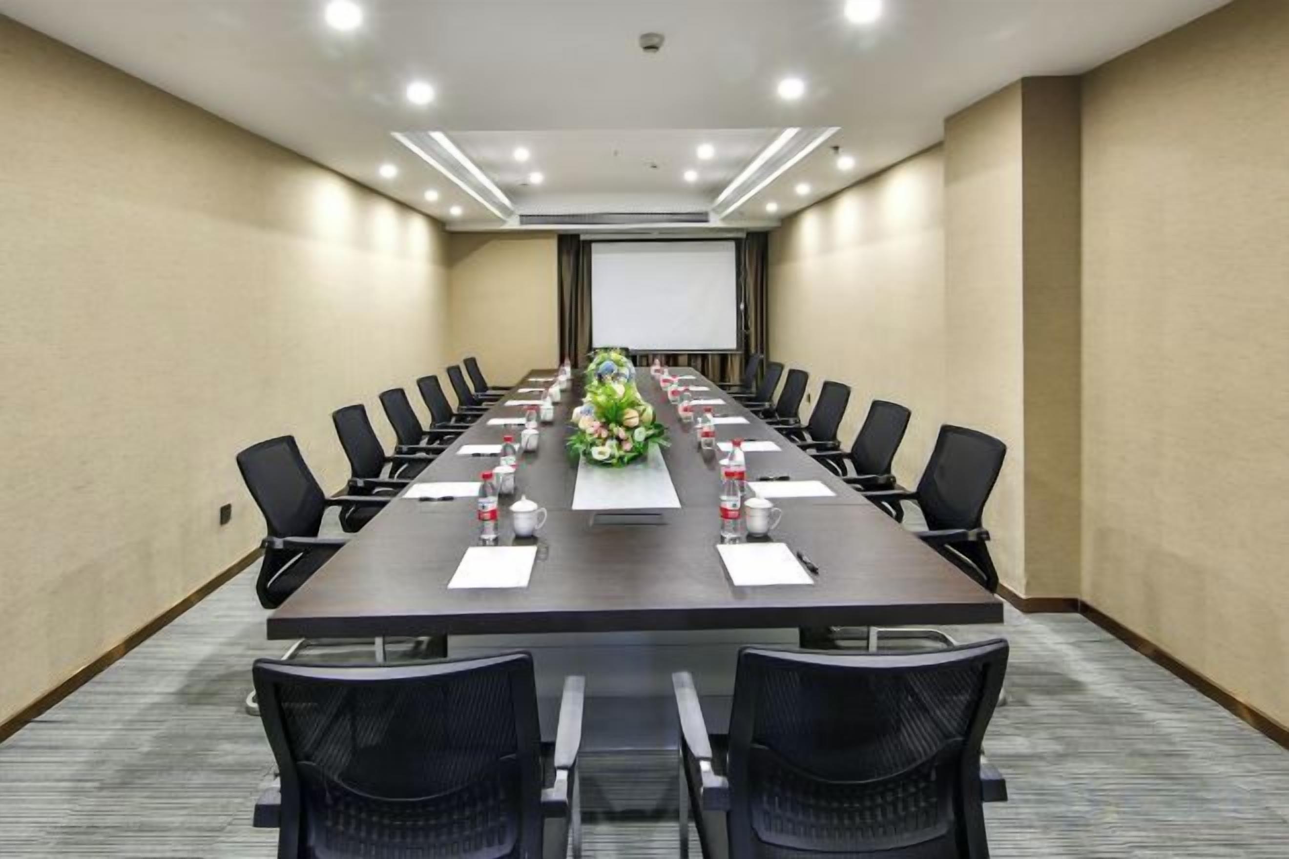 A conference room with a long table, chairs, a projector screen, and a floral centerpiece.